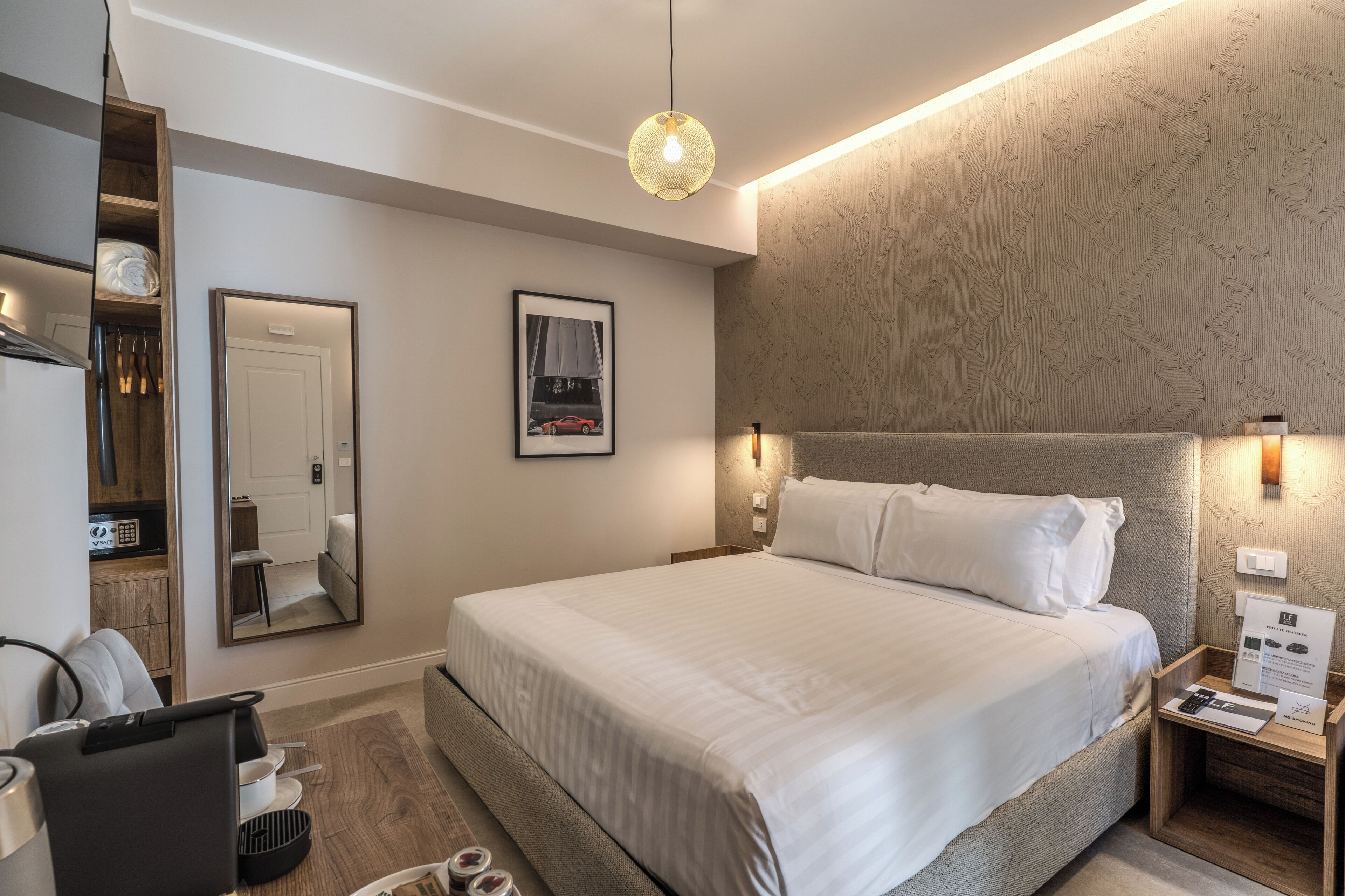 A modern hotel room with a large bed, a bedside table with a lamp, a mirror, and a framed picture on the wall.