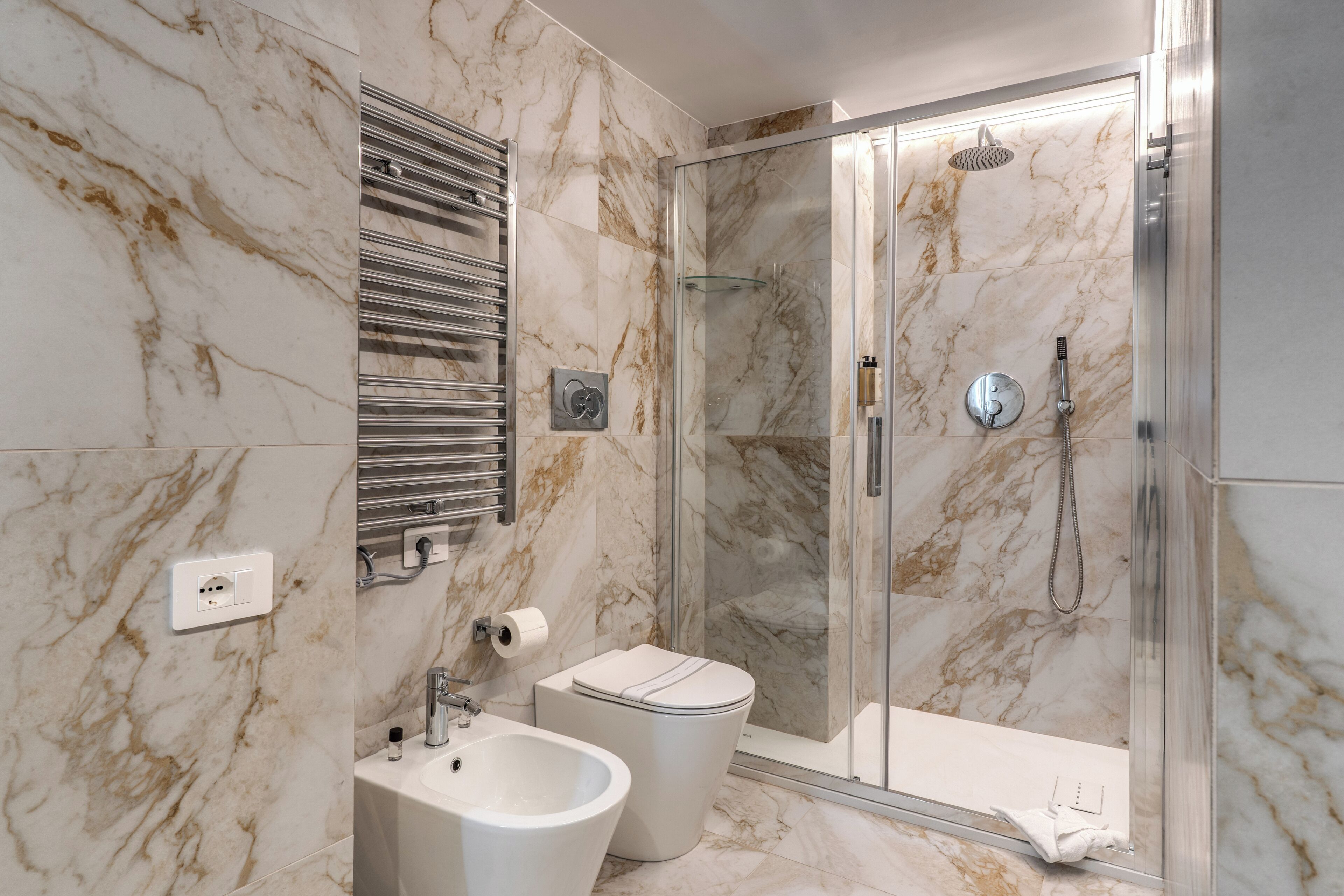 A bathroom with a marble wall, a glass shower enclosure, a bidet, and a toilet.