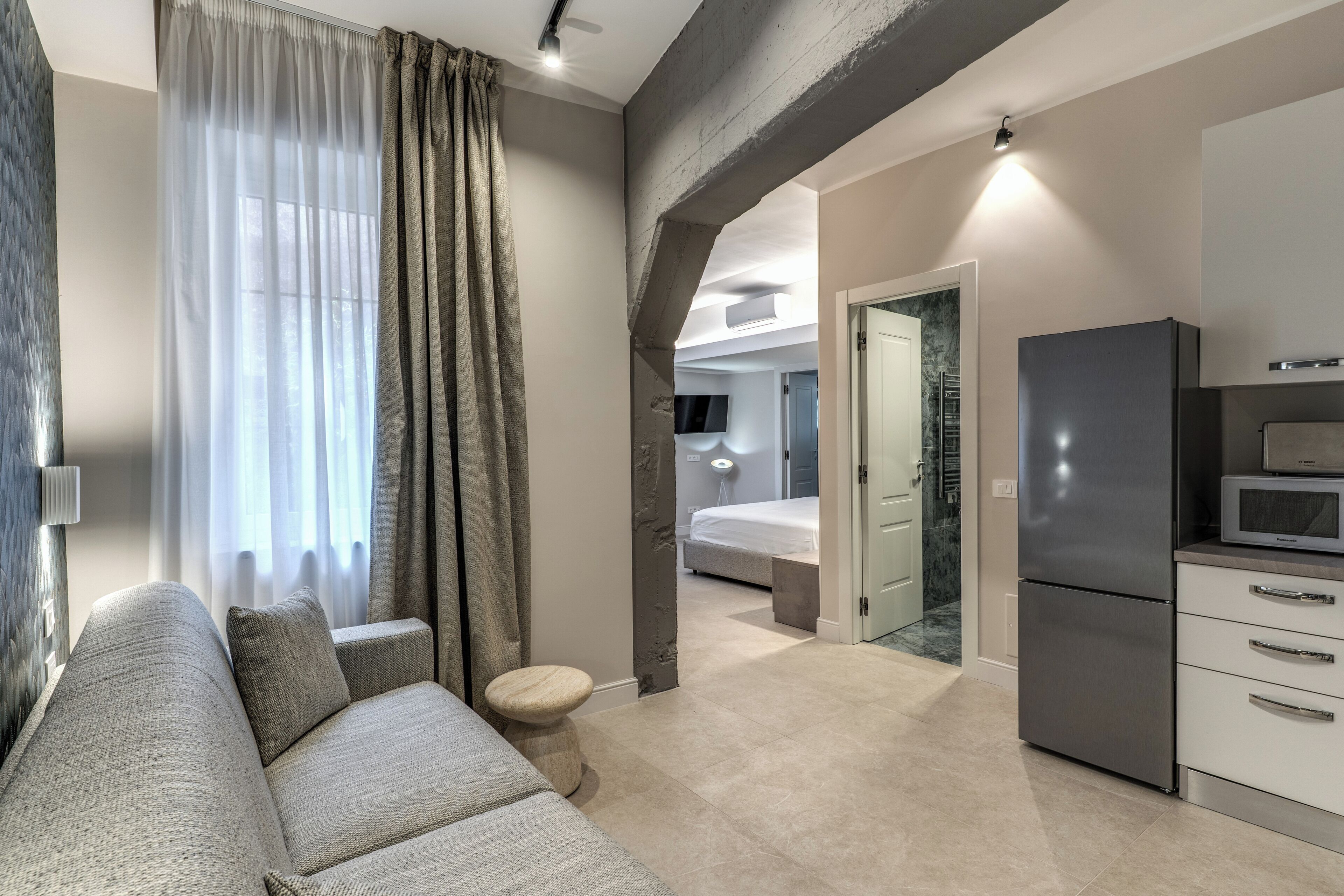 A modern living room with a sofa, a small stool, a kitchen area with a refrigerator, and a bedroom with a bed and a TV.