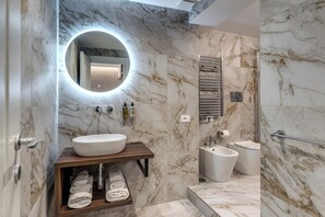 Un bagno moderno con parete in marmo, uno specchio rotondo, una toeletta in legno, una vasca da bagno e un water.