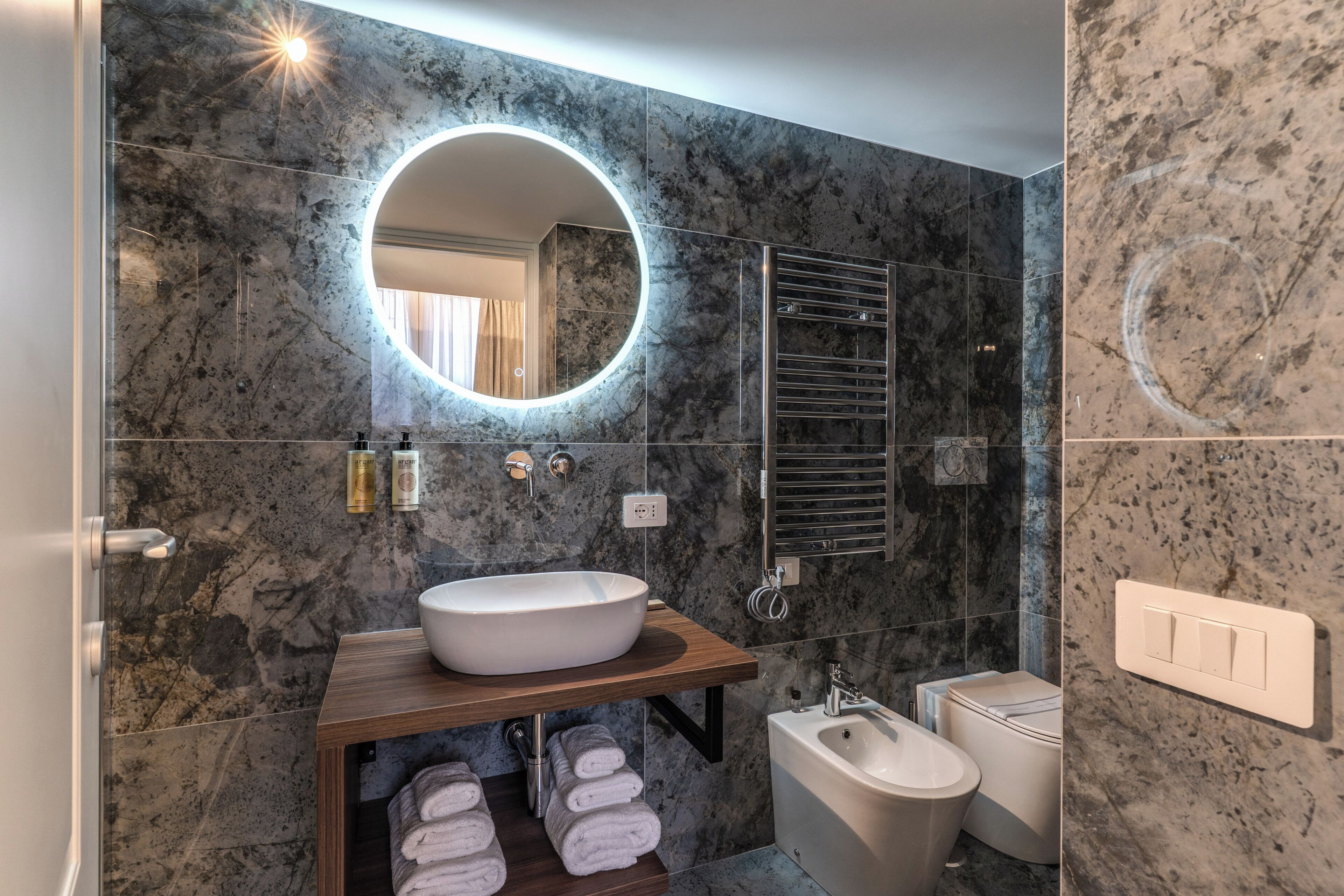 A bathroom with a marble wall, a round mirror, a sink, a toilet, and a towel rack.