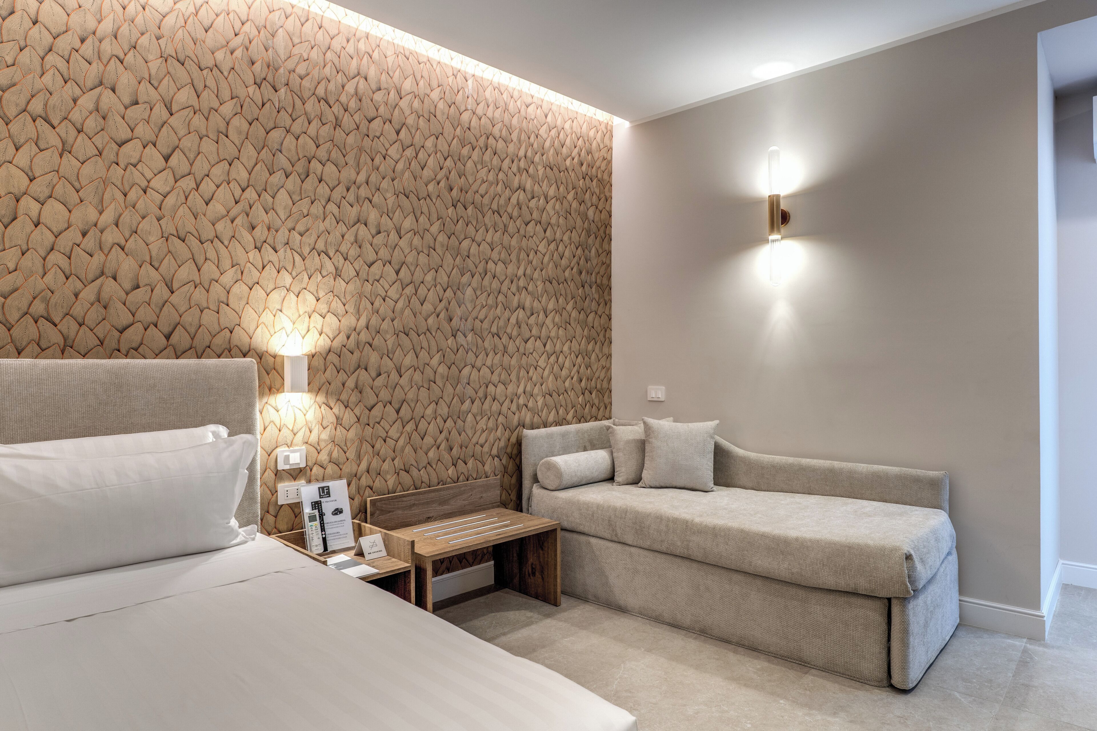 Habitación de hotel con pared texturizada, cama, sofá, mesita de noche con lámpara y luz empotrada en la pared.
