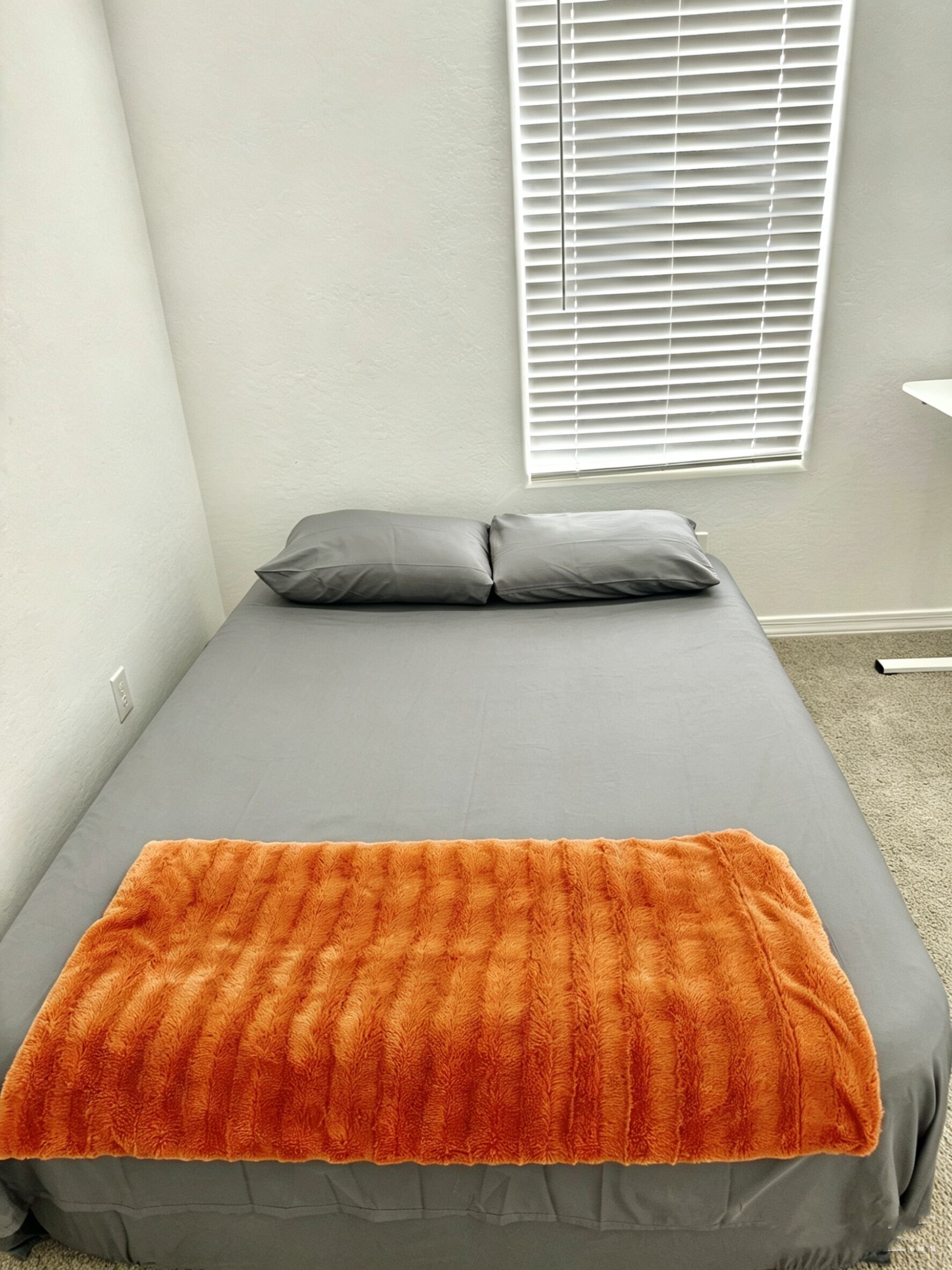 A small, neatly made bed with grey bedding and an orange blanket, a white wall, and a window with blinds.