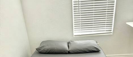 A small, neatly made bed with grey bedding and an orange blanket, a white wall, and a window with blinds.