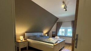 Un dormitorio con techo inclinado, una cama con almohadas y una colcha, una mesita de noche con lámpara, y una ventana con cortinas.