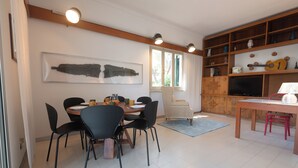 Ein Essbereich mit einem runden Tisch, schwarzen Stühlen, einem gemusterten Sessel und einem Fernseher auf einem Holzboden.