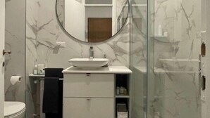 Un baño con pared de mármol, un espejo redondo, un lavamanos, un inodoro y una cabina de ducha de vidrio.