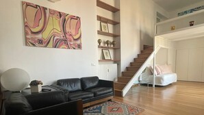 Una sala de estar con un sofá de cuero negro, un cuadro abstracto colorido, una escalera de madera y un sofá blanco al fondo.