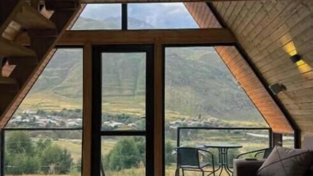 A room with a sloped ceiling, large windows, a wooden floor, a sofa, and a small table. - Your Peaceful Retreat in Kazbegi – Chalet with Stunning Views & Warm Charm (HHWF+JH, Sioni, Georgia)