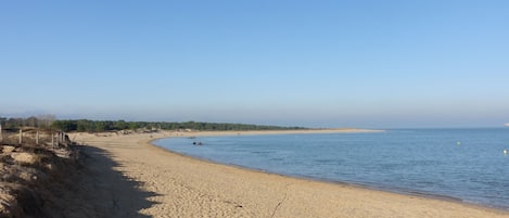 A wide sandy beach with gentle waves, a clear sky, and a distant shoreline with greenery.