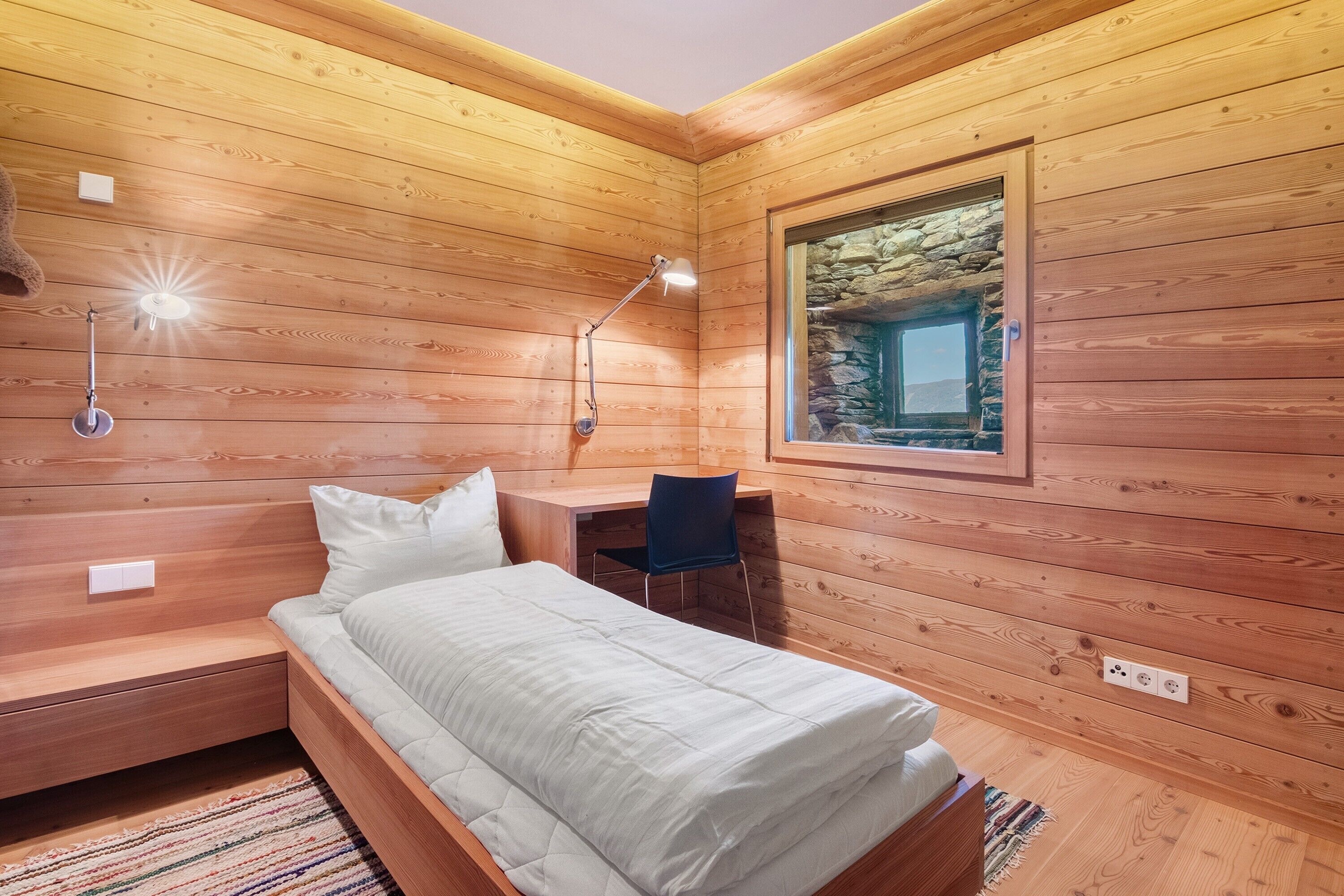 Una stanza con pannelli in legno, un letto, una scrivania, una sedia e vista su un muro in pietra.