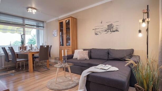En moderne stue med en grå sofa, et spisebord med stole, et glasvitrineskab og et stort vindue.