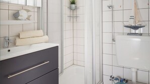 Et moderne badeværelse med en glasbrusekabine, et hvidt håndvaskarrangement og en mørk kommode med en lille plante i potte.