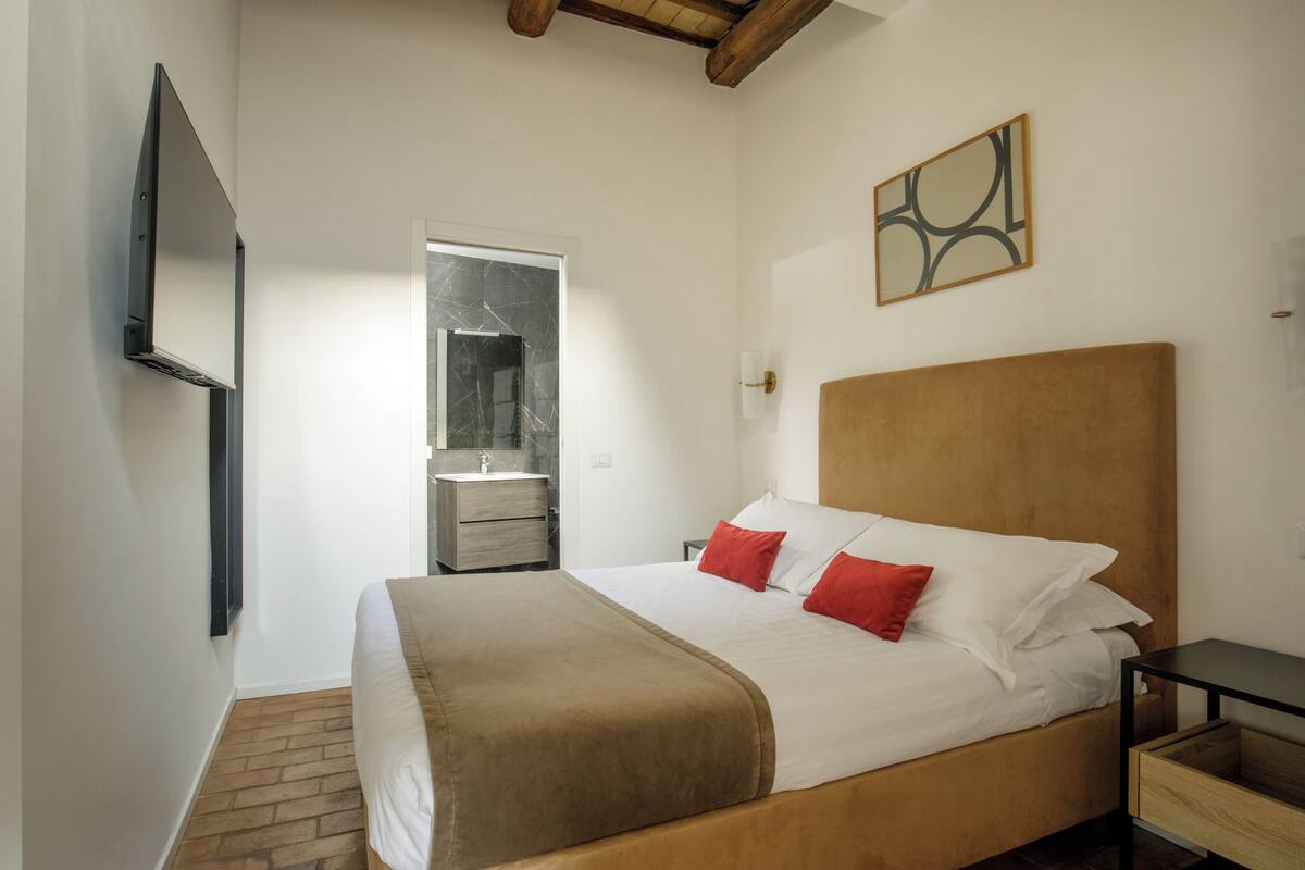 A bedroom with a bed, a nightstand, a TV mounted on the wall, and a bathroom visible through an open door.