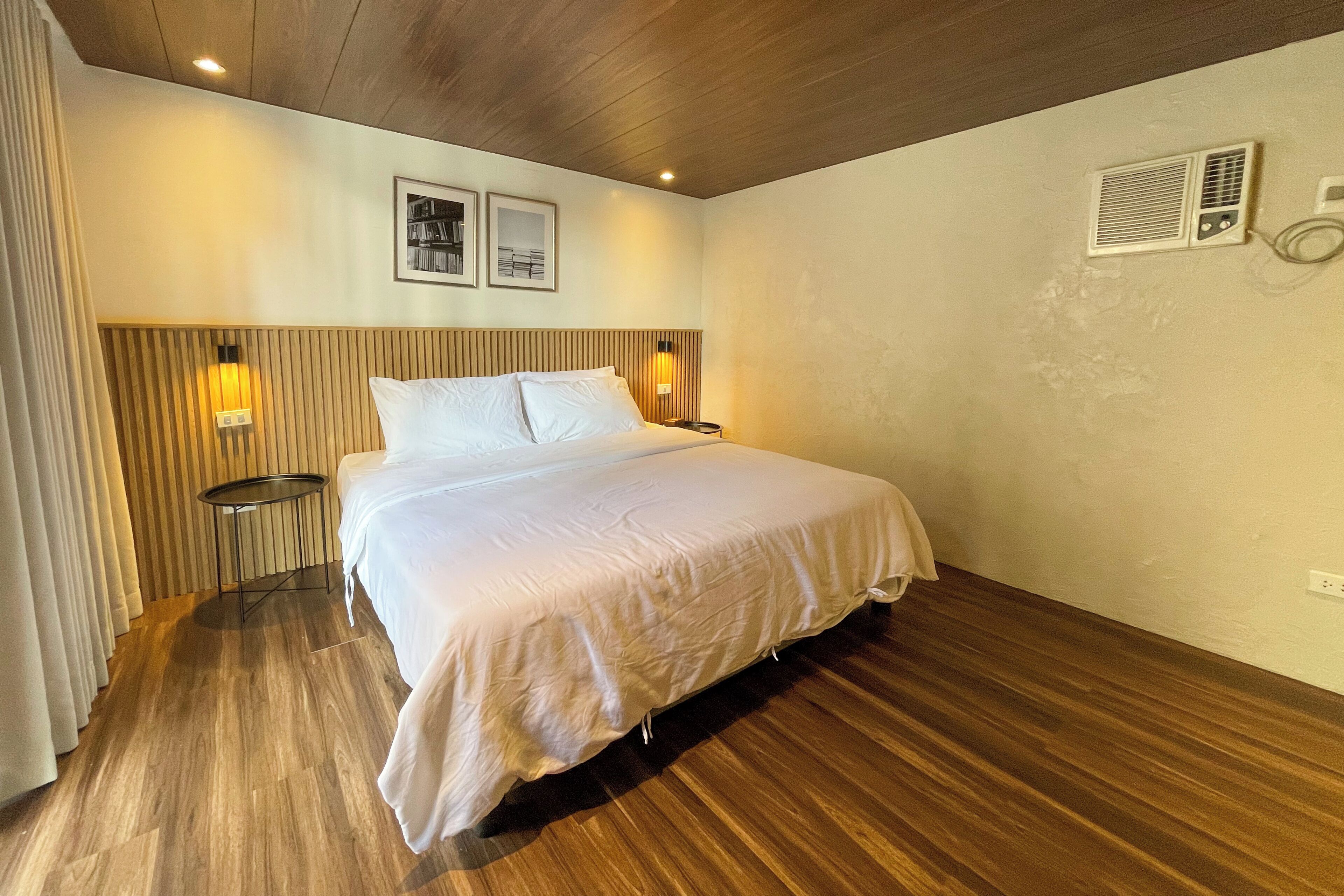 A hotel room with a wooden ceiling, a bed with white bedding, a round side table, a wall-mounted heater, and two framed pictures.