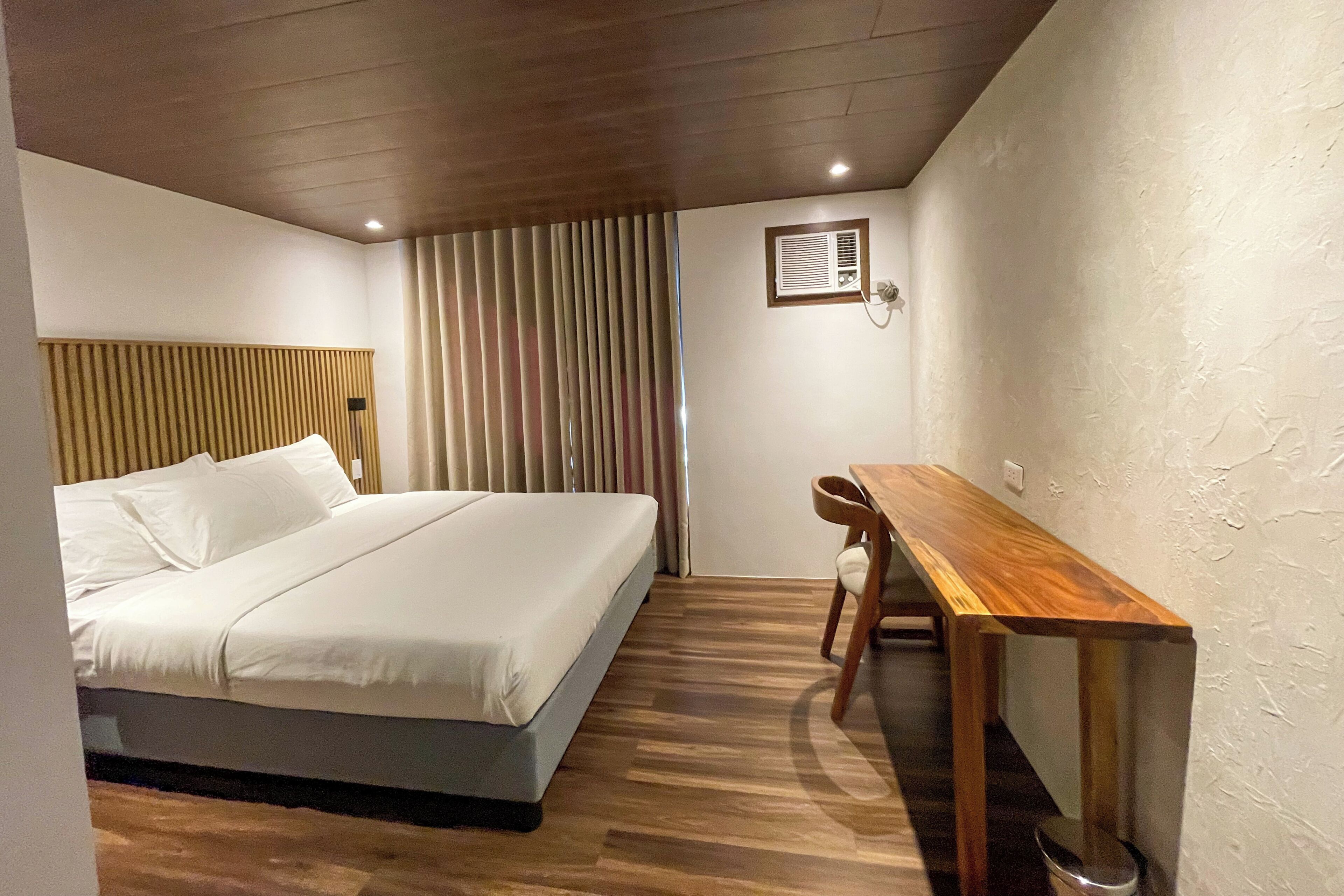 A bedroom with a bed, a wooden desk, a chair, a small table, a wall-mounted air conditioning unit, and a window with blinds.
