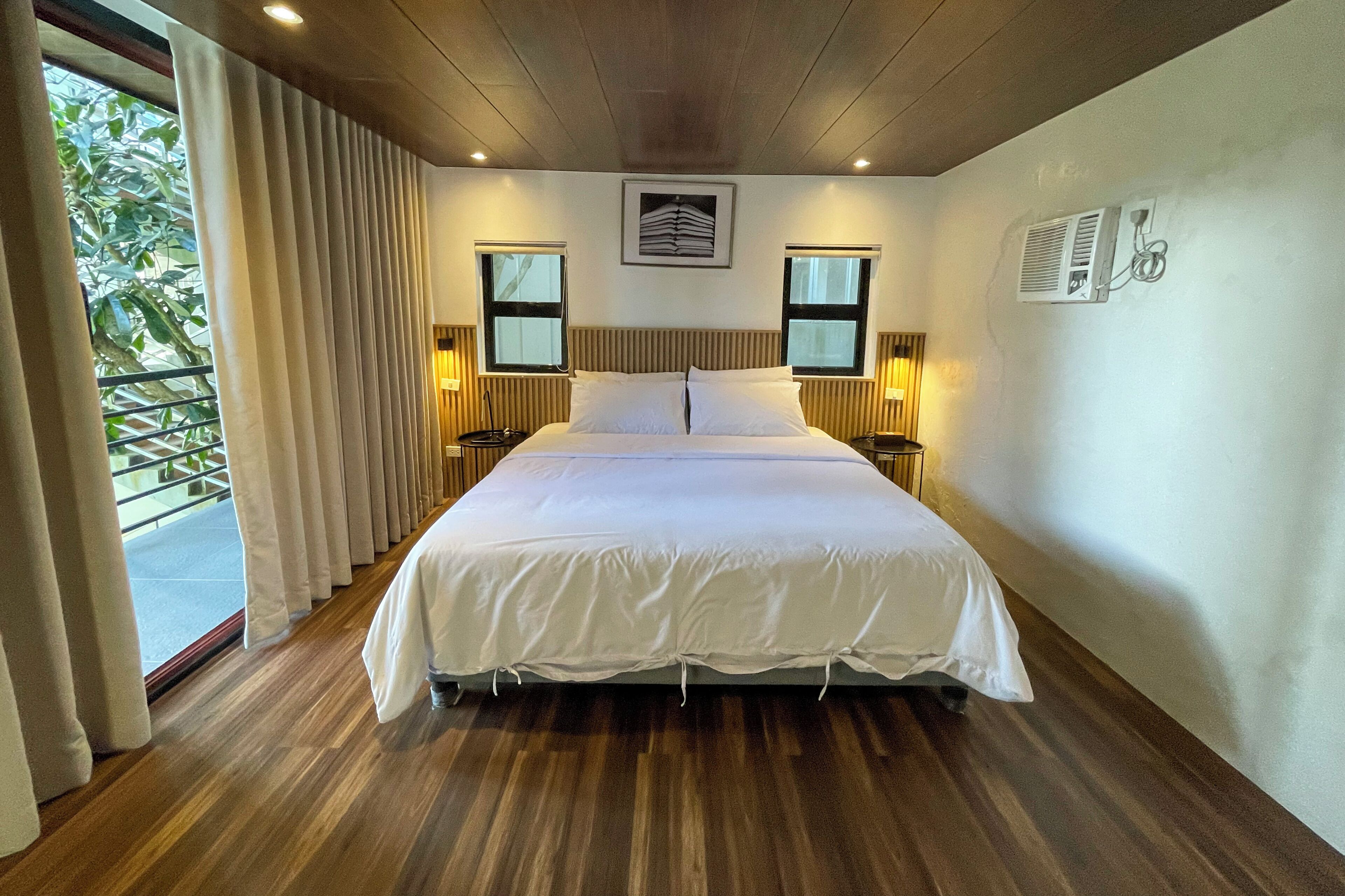 A hotel room with a wooden ceiling, a large bed with white bedding, two bedside lamps, and a window with curtains.