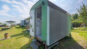 A green mobile home with a door open, a small step leading up to the entrance, and a grassy yard with a metal chair and a trash bin. - Bauwagen 'Natur Pur/No.7' with Private Terrace and Panoramic Countryside Views (Ötisheim)