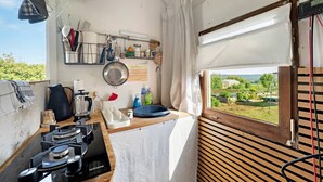 A compact kitchen with a gas stove, hanging utensils, and a window with a view of greenery. - Bauwagen 'Natur Pur/No.7' with Private Terrace and Panoramic Countryside Views (Ötisheim)