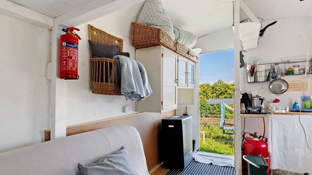 A compact room with a bed, a fire extinguisher, a small kitchenette, and a view of greenery outside. - Bauwagen 'Natur Pur/No.7' with Private Terrace and Panoramic Countryside Views (Ötisheim)