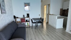 Ein modernes Wohnzimmer mit grauem Sofa, einem Esstisch mit Stühlen, einer Küchenzeile mit weißer Arbeitsplatte und einem gerahmten Bild an der Wand.