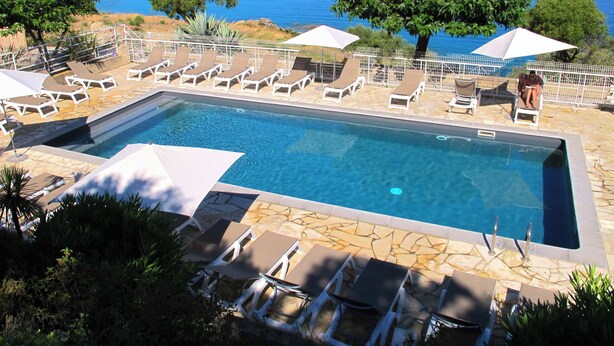 Ein rechteckiger Pool mit Liegestühlen und Sonnenschirmen, umgeben von Steinplatten, mit Blick auf eine Küstenlandschaft und klares blaues Wasser sowie Land in der Ferne.
