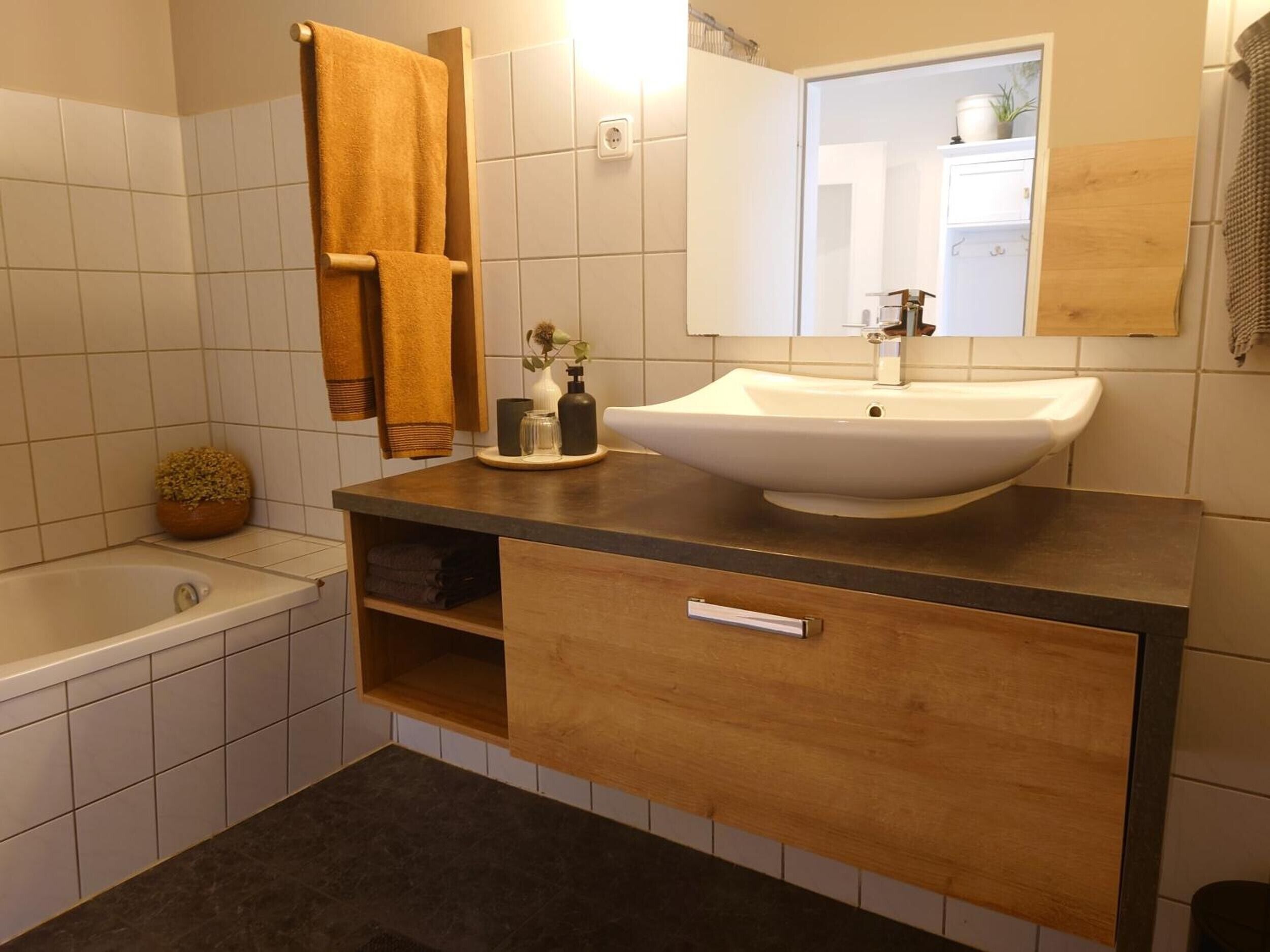 Ein Badezimmer mit einem weißen Waschbecken, einem Holz-Badmöbel, einem Handtuchhalter mit Handtüchern, einer kleinen Topfpflanze und einem Spiegel.