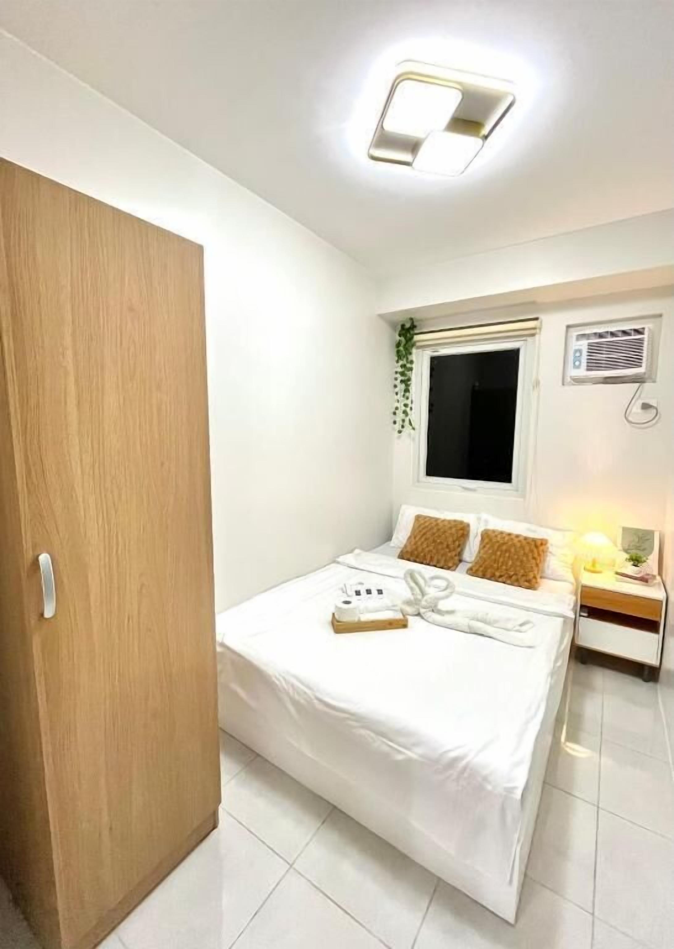 A hotel room with a bed, bedside table, window, and a ceiling light fixture.