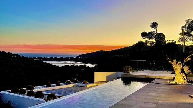 Una piscina infinita con vistas al océano y colinas al atardecer.