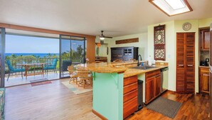 A kitchen with a large island, a dining area with chairs, a view of the ocean, and a ceiling fan. - Sunset View at Kahaluu Bay Villas (Kailua-Kona)