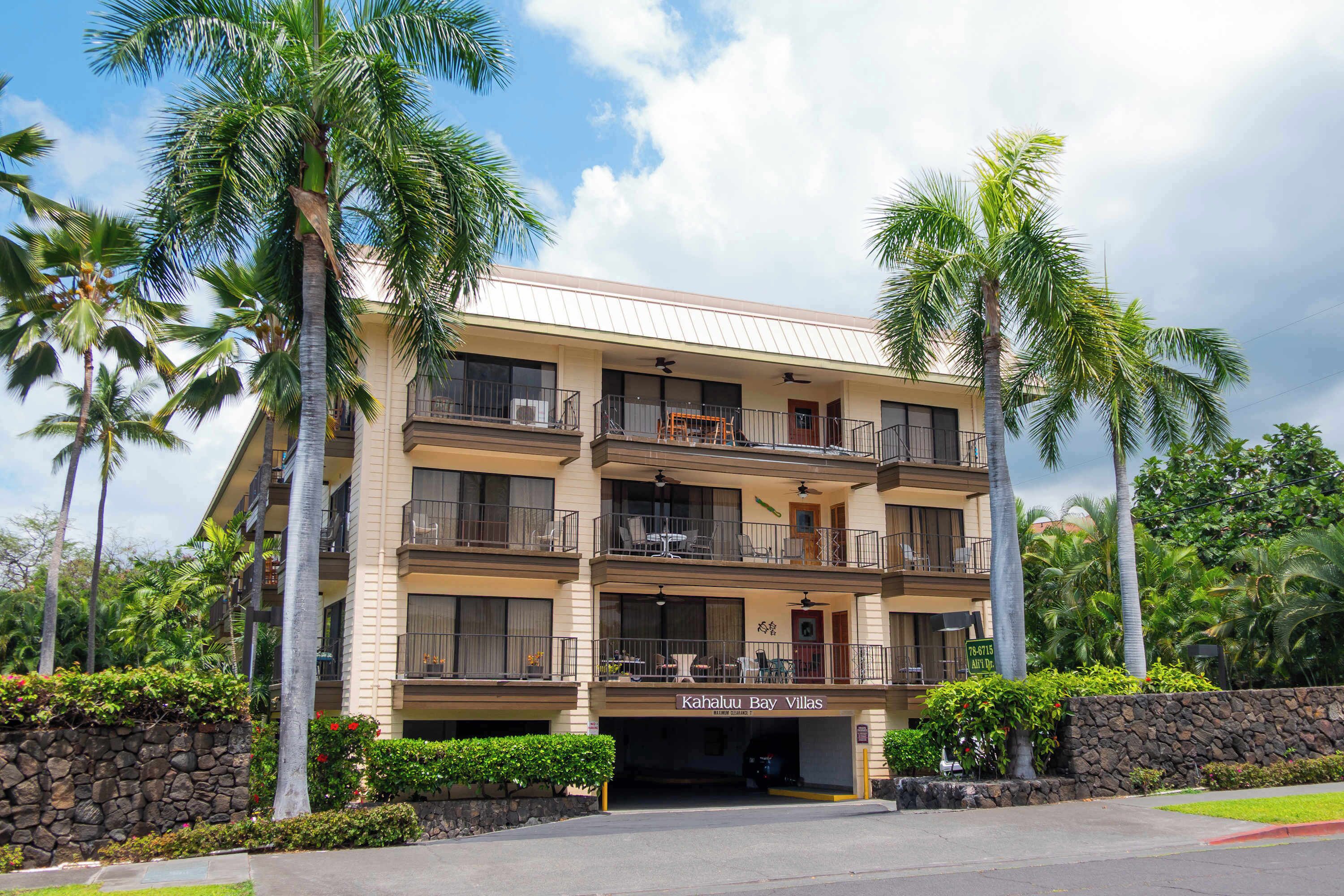 A multi-story hotel with balconies, a stone wall, and palm trees.