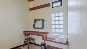 A small bathroom with a wooden bench, a trash can, a towel rack, and a window with a decorative glass pattern. - Kampung Bingo Glamping & Resto (Baturiti)