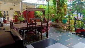 A covered patio area with a table and chairs, surrounded by potted plants and a stationary exercise bike. - Homestay Urmil Basant (Jaipur)