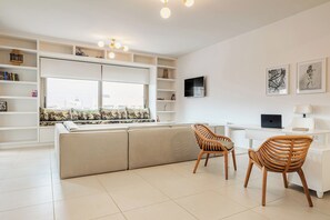 Ein modernes Wohnzimmer mit einer Couch, zwei Holzstühlen, einem weißen Schreibtisch mit einem Laptop, einem Fernseher und einem Bücherregal.