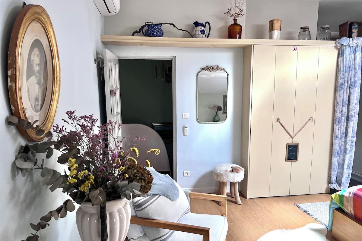 Una habitación con cama, una silla, un taburete pequeño, un jarrón blanco con flores, un espejo, una estantería con objetos decorativos y un armario.