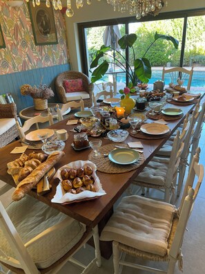 Una mesa de comedor dispuesta con diversos alimentos y bebidas, rodeada de sillas de mimbre y vista a una piscina.