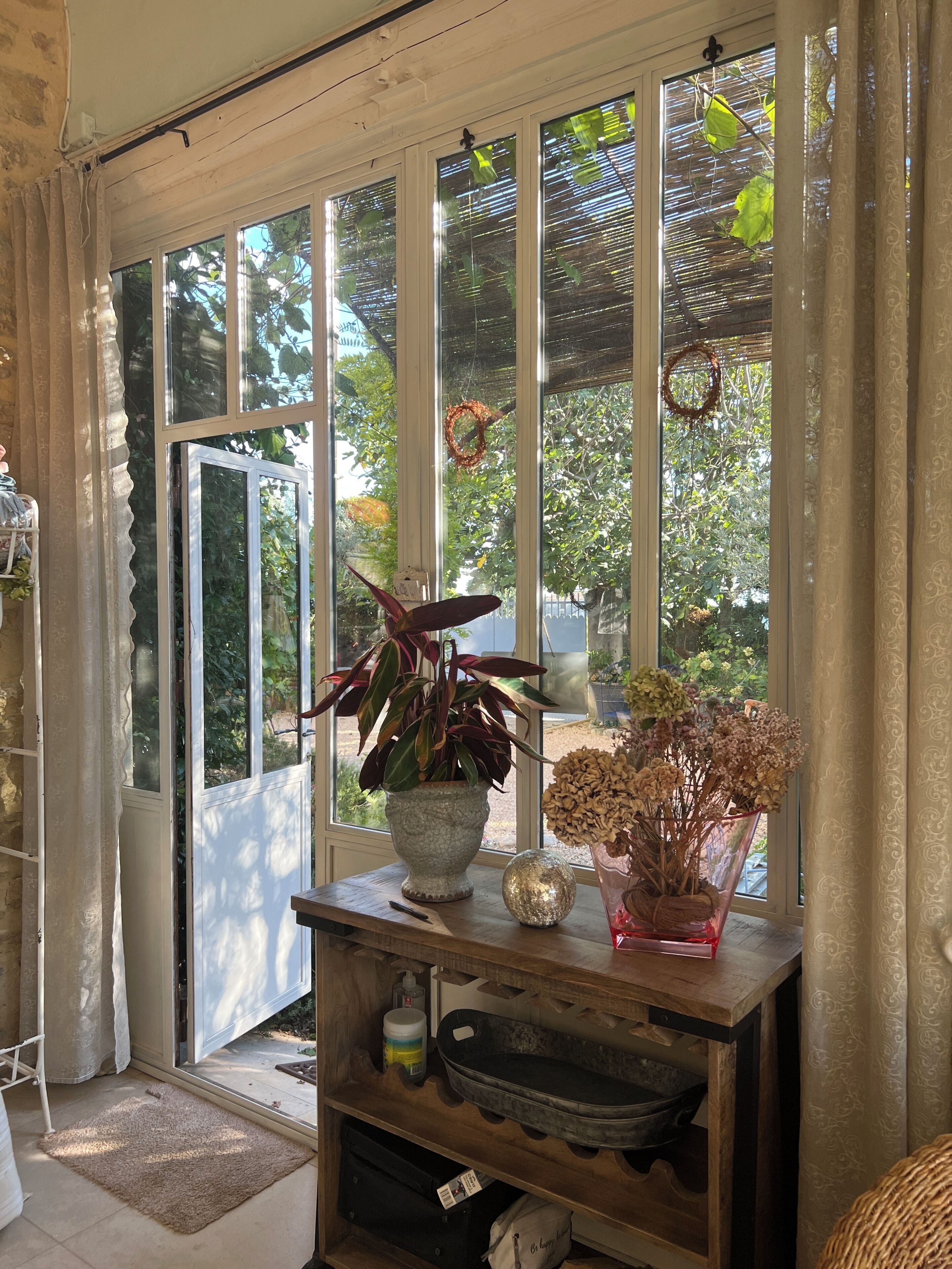 Une pièce dotée d’une grande porte-fenêtre coulissante donnant sur un jardin, d’une table en bois sur laquelle est posée une plante en pot, et d’une chaise en osier.