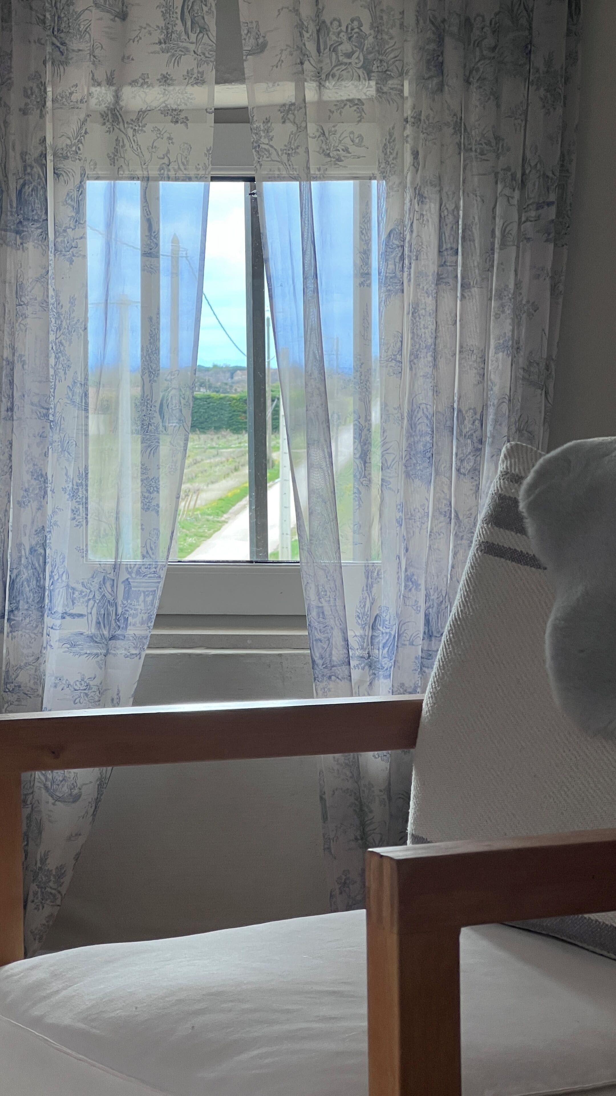 Una habitación con una ventana cubierta por cortinas florales, una silla de madera y un cojín blanco.
