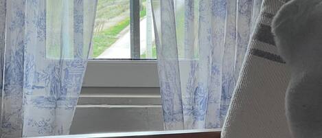 Una habitación con una ventana cubierta por cortinas florales, una silla de madera y un cojín blanco.