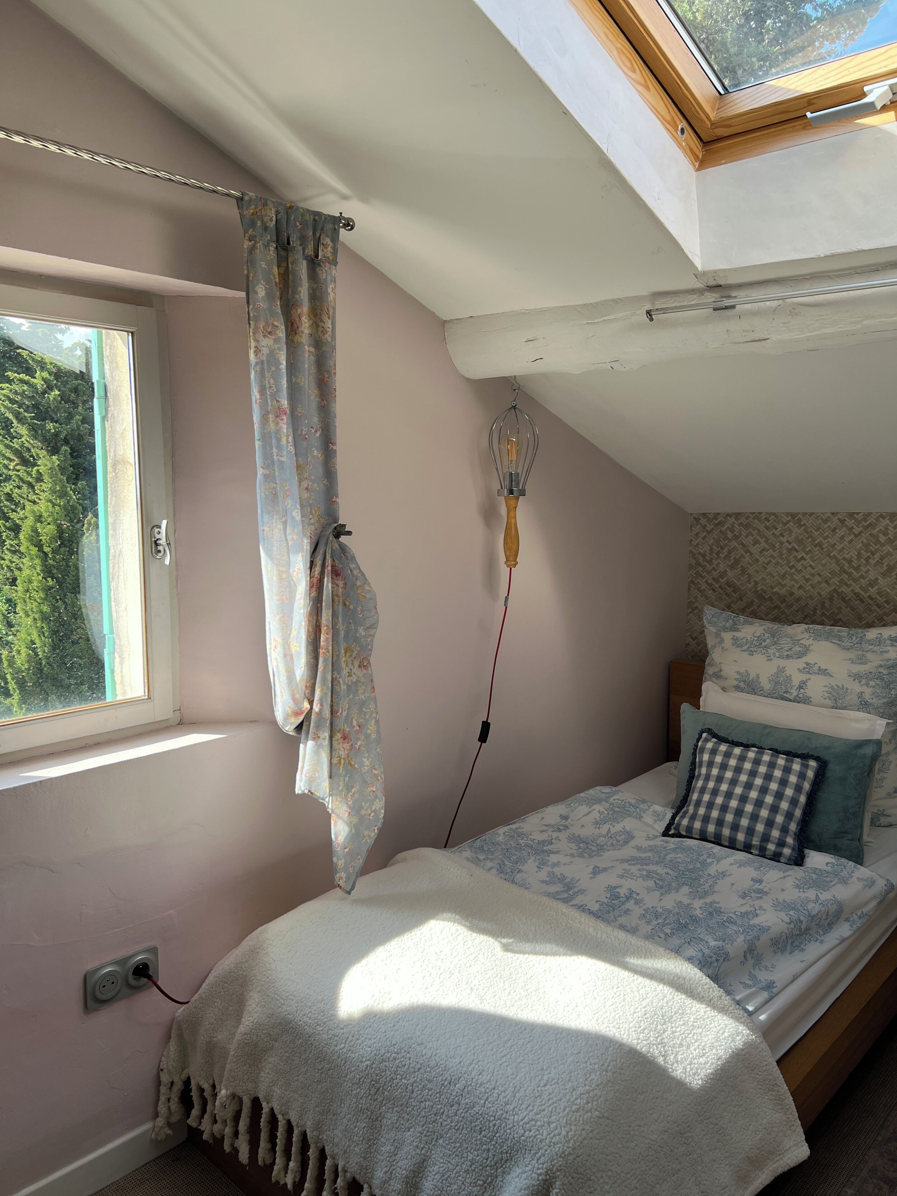 Een slaapkamer met dakraam, een bed met beddengoed met dessin, een hanglamp en een raam met gordijnen.