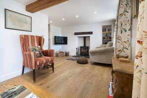A living room with a wooden floor, a striped armchair, a sofa, a television, a fireplace, and a bookshelf. - Garden Cottage -  Cosy Cottage in Brigsteer (Brigsteer)