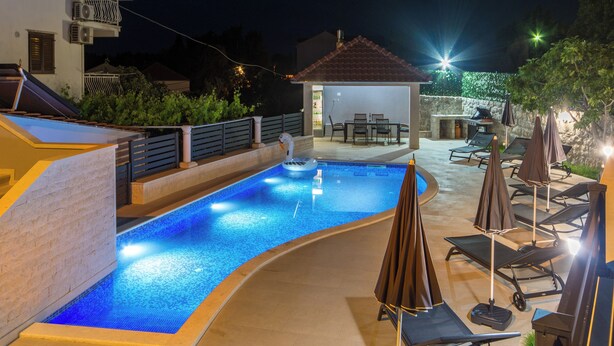 Une piscine à bord incurvé, entourée de chaises longues et de parasols, et un espace barbecue à l’arrière-plan.