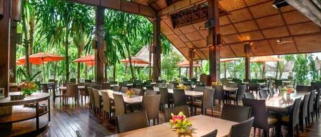 An indoor dining area with tables and chairs, a thatched roof, and large windows.