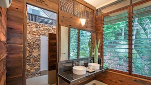 A bathroom with wooden walls, a stone accent wall, a large mirror, and a view of greenery through the window. - Rawa Island Resort (Mersing)