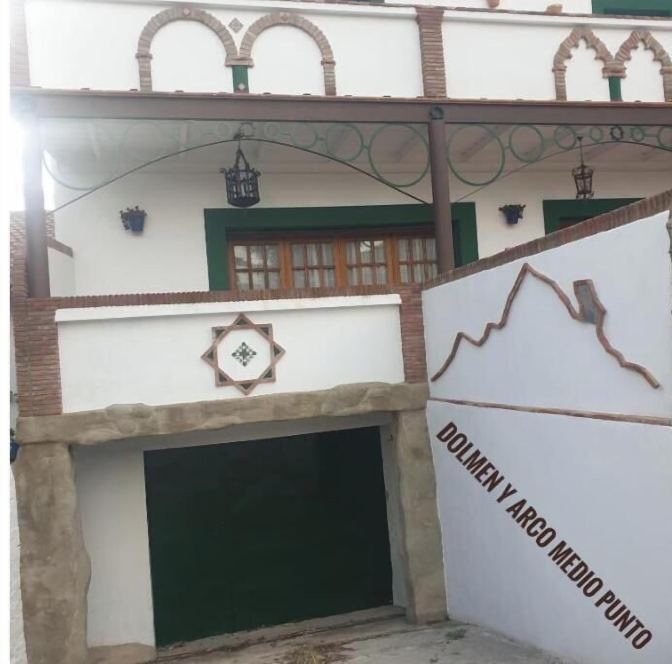 A building with a green window, a decorative wall, and a sign that reads 'DOLMEN Y ARCO MEDIO PUNTO'.