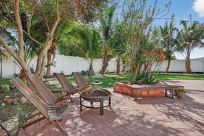 A patio with lounge chairs, a fire pit, and a garden with palm trees and a white fence. - Coconut Oasis Near Beach (Lauderdale-by-the-Sea)
