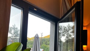 Une pièce dotée d’une grande fenêtre donnant sur un patio aménagé avec du mobilier d’extérieur, et offrant une vue sur les arbres et les collines.