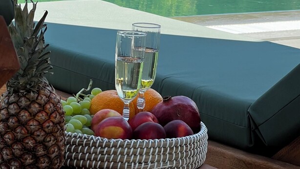 Une table placée au bord de la piscine, sur laquelle se trouve un panier de fruits contenant des ananas, des raisins, des pêches et deux verres d’eau.