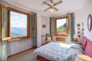 Um quarto com uma cama grande, um ventilador de teto, uma janela com vista para as montanhas e uma mesa de cabeceira com abajur.