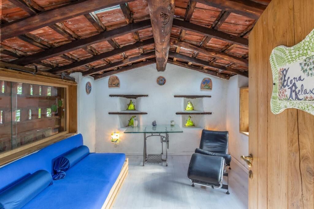 Een kamer met een blauwe bank, een zwarte stoel, een glazen tafel en houten planken met decoratieve objecten.